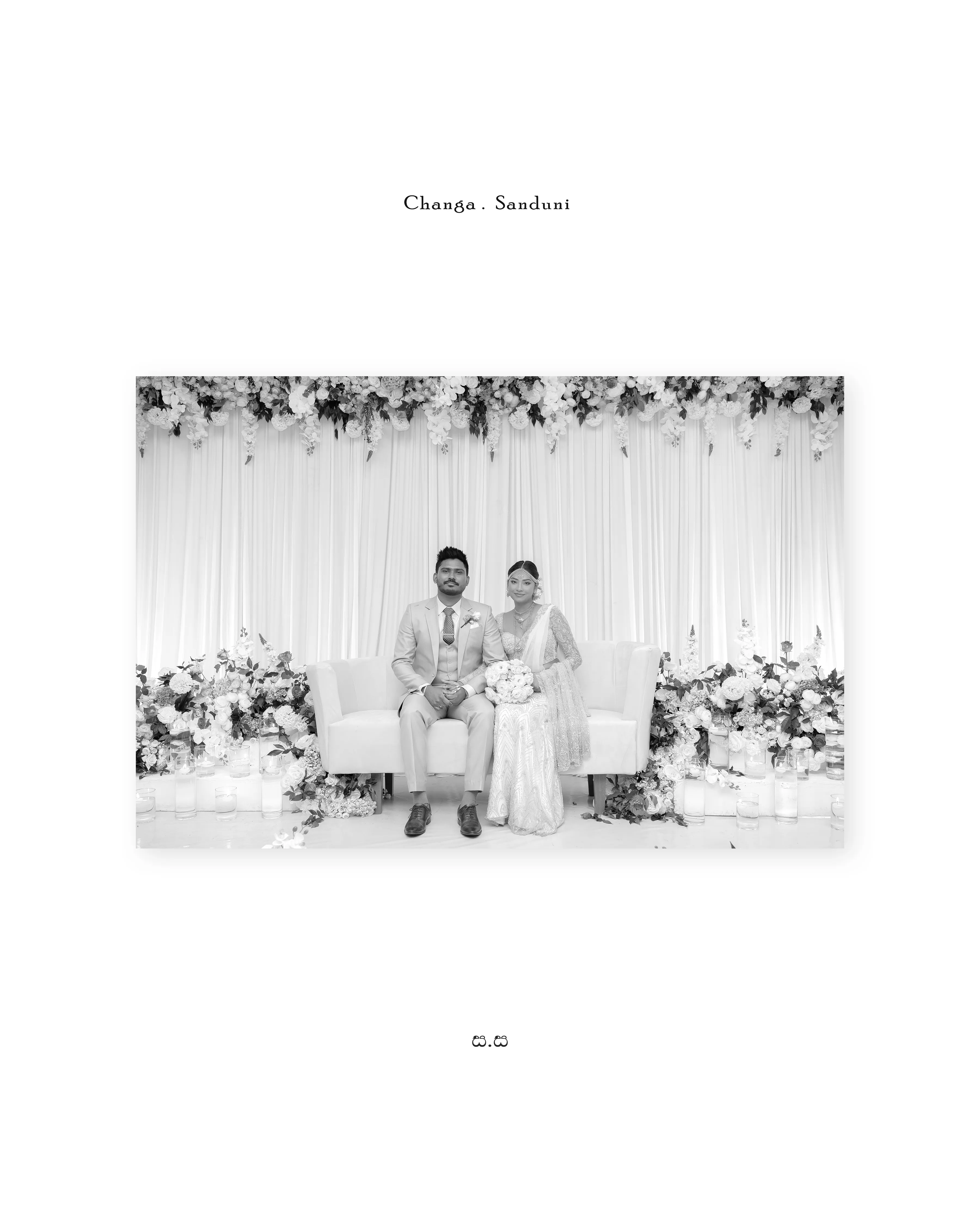 The newlyweds sit together beneath a white floral backdrop in black and white.