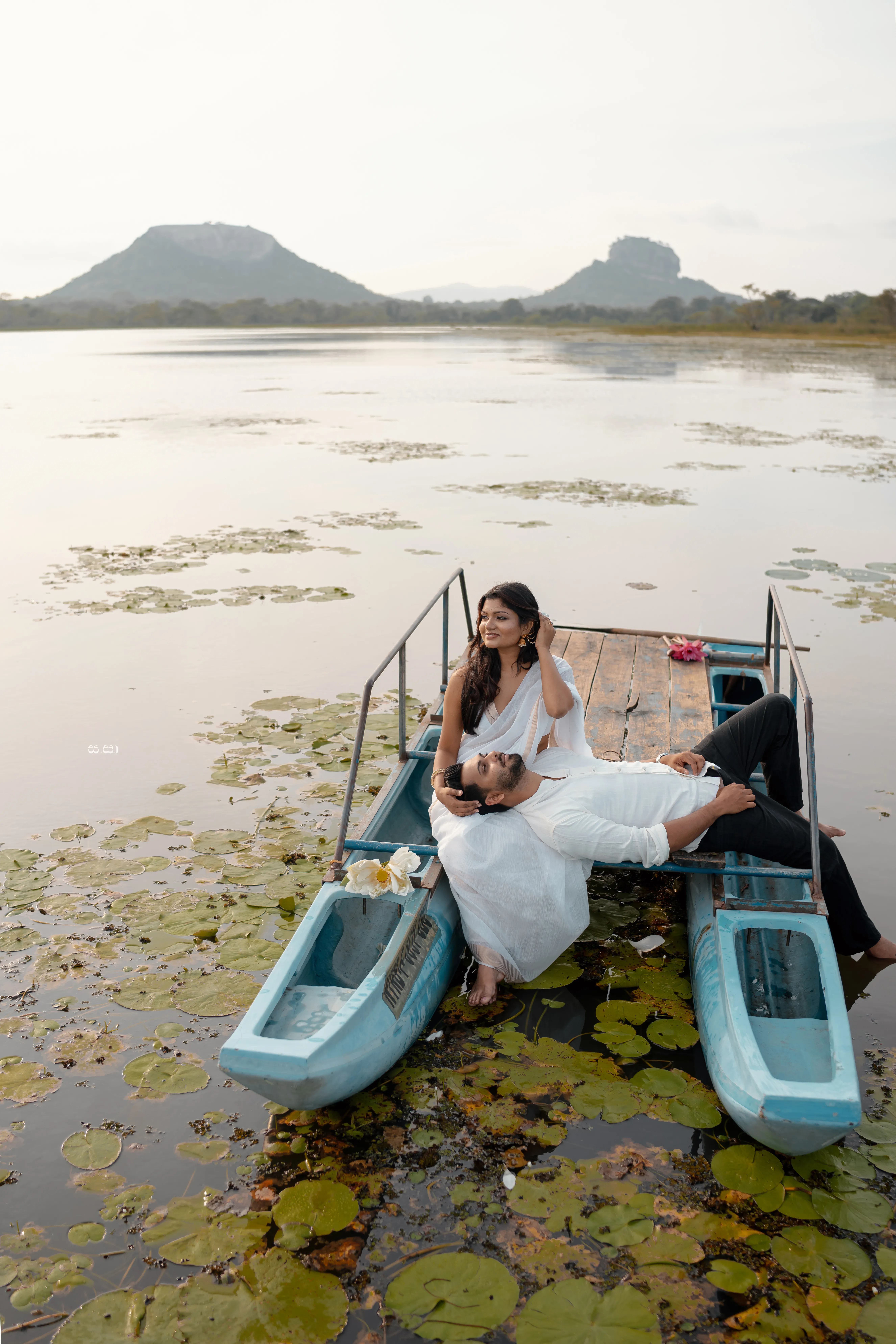 The couple sits close in a blue boat drifting through a lotus-covered lake.