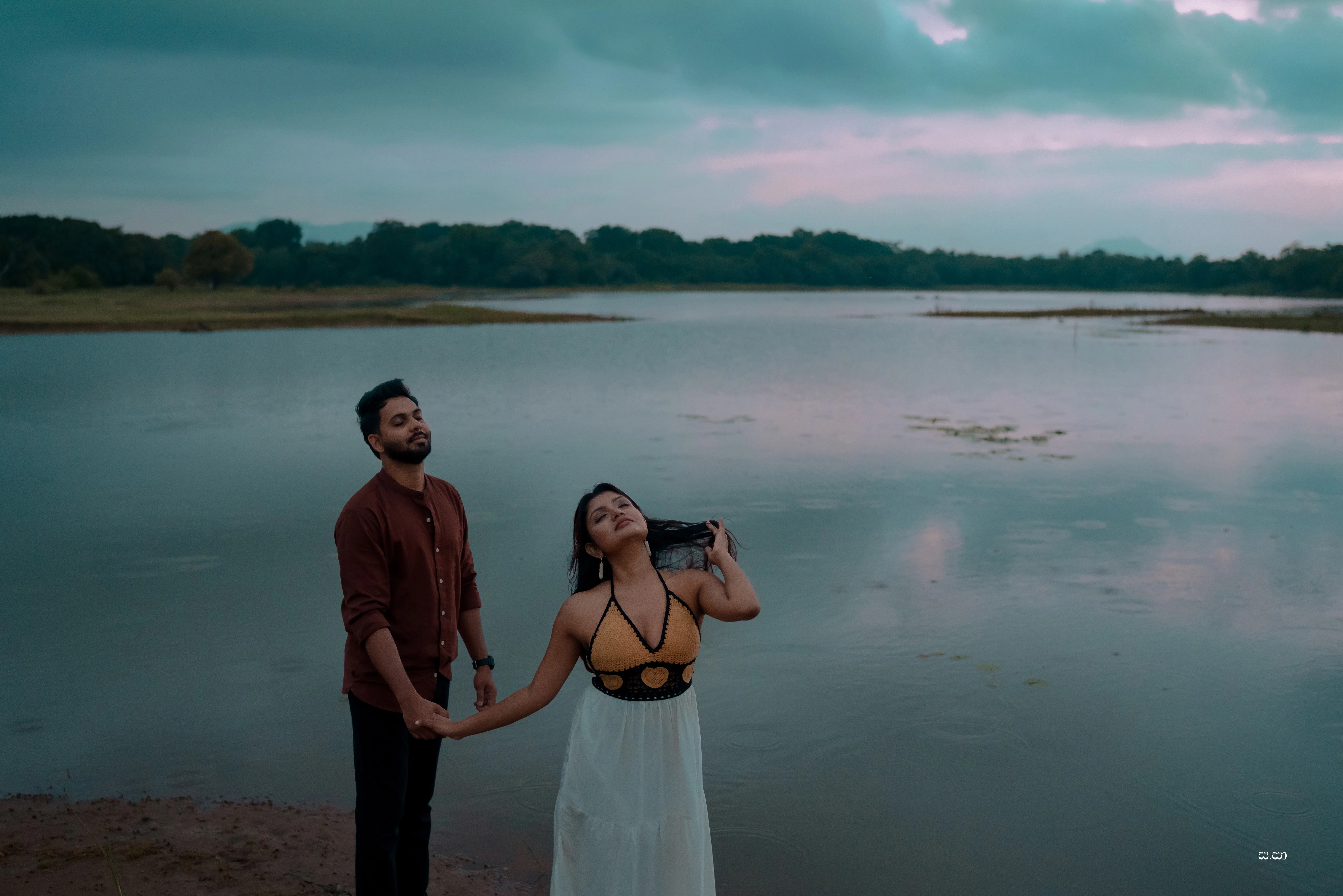 Lakshani laughs beside Charith under a vivid blue-grey lakeside sky.