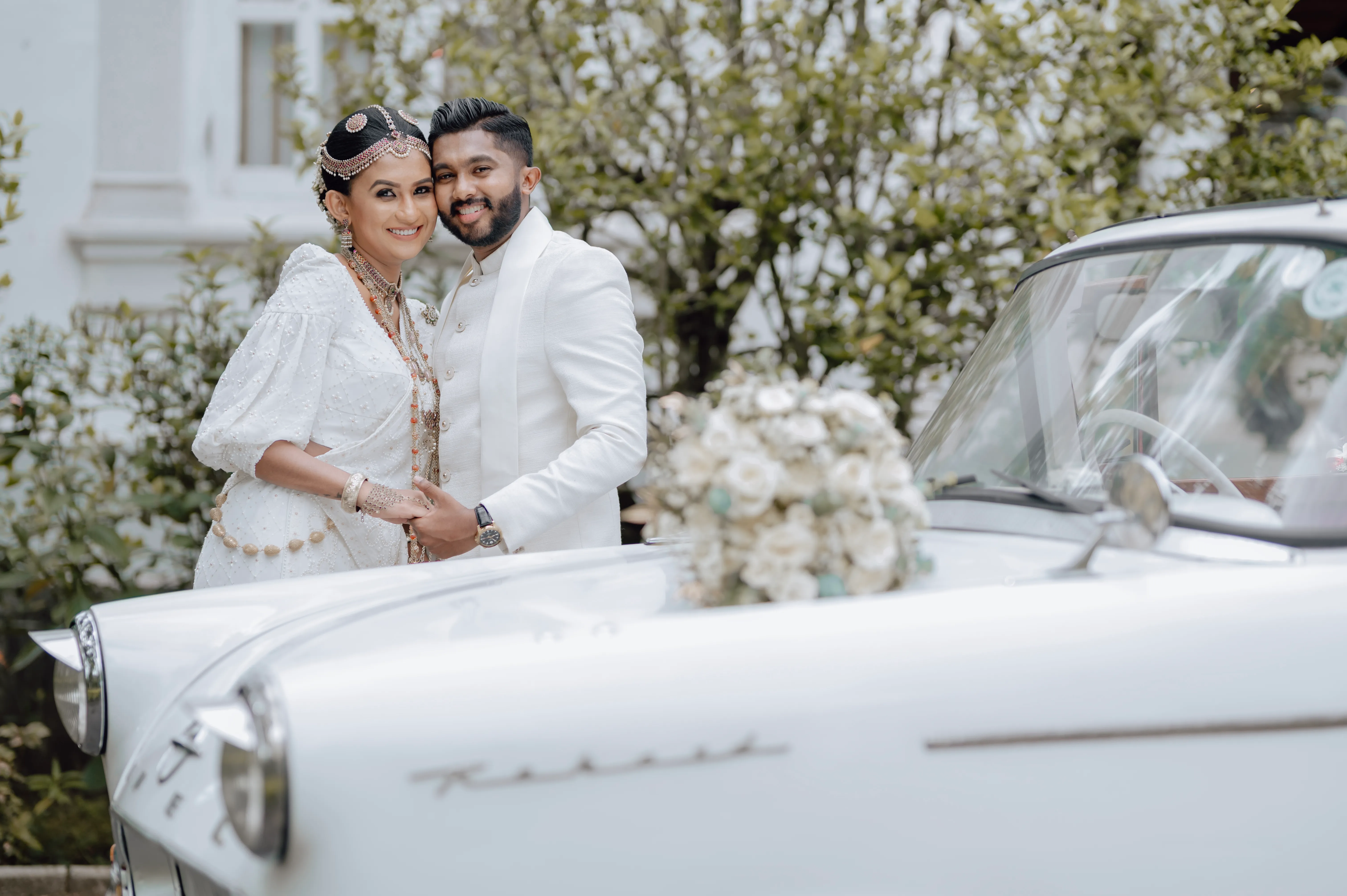 The couple embraces beside a white vintage car covered with pale flowers.