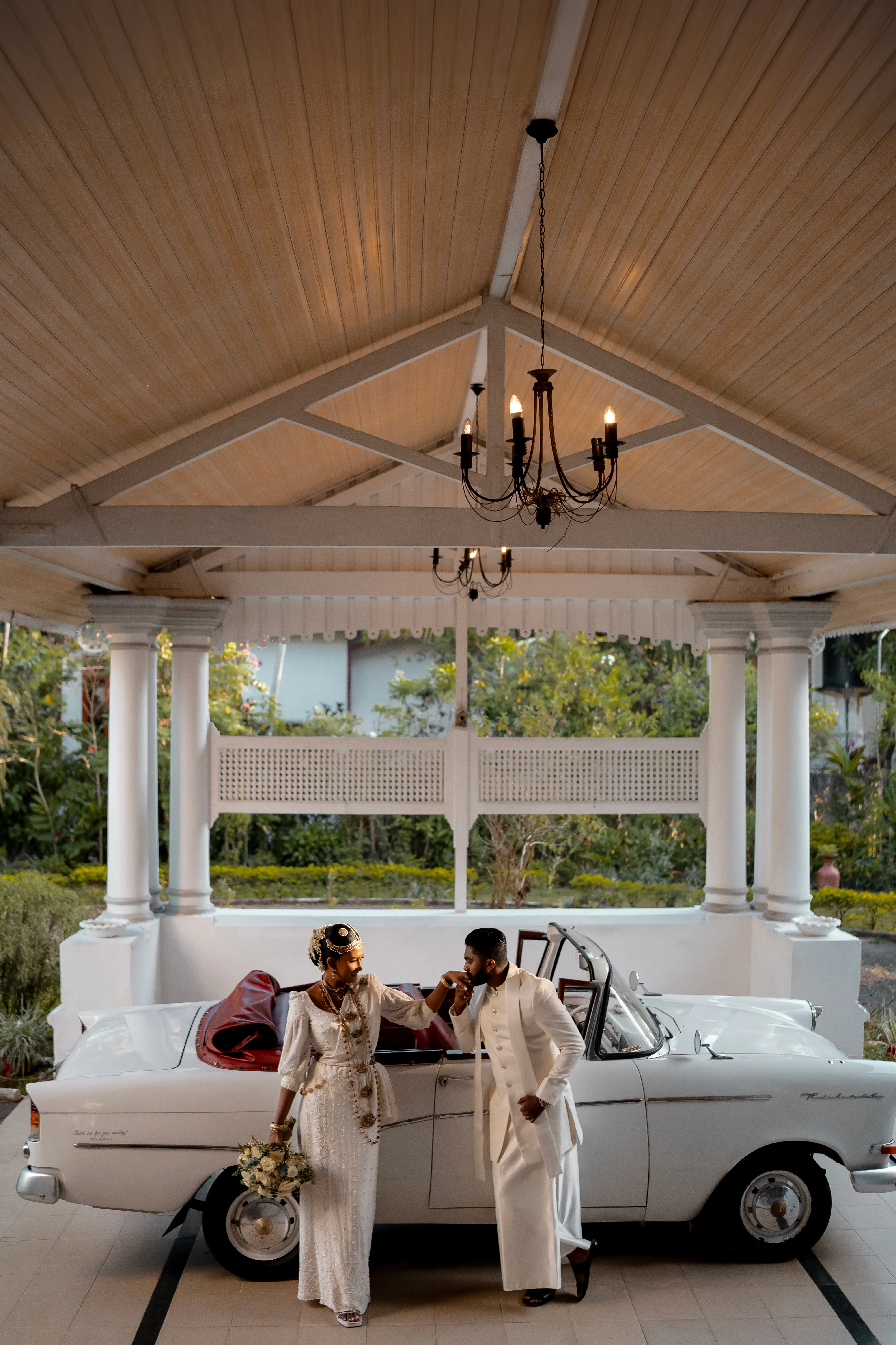 The couple stands beside the vintage car beneath a white garden portico.
