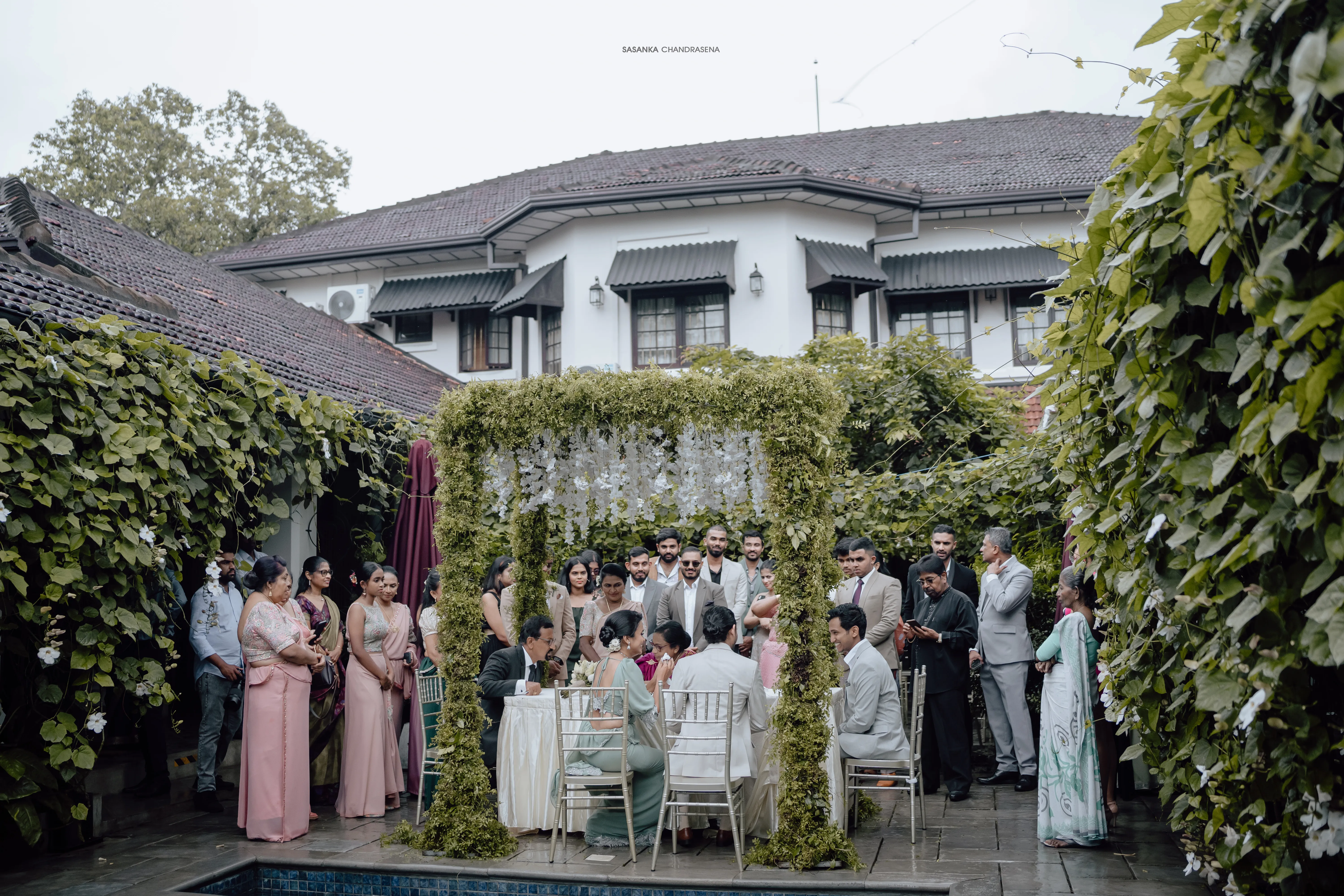 Family and friends gather beneath greenery for the outdoor engagement ceremony.