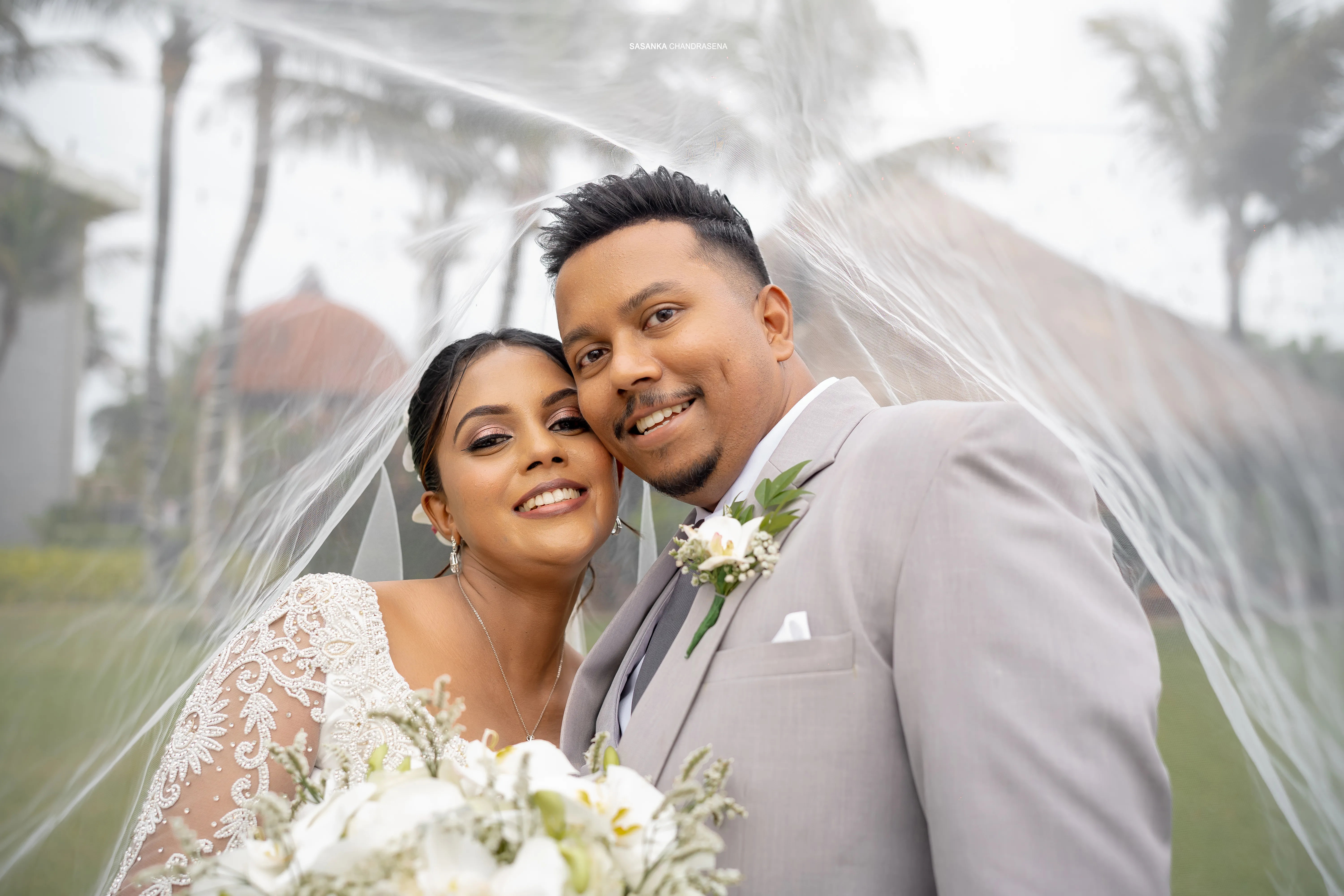 The couple smiles together beneath palms with white flowers in the foreground.