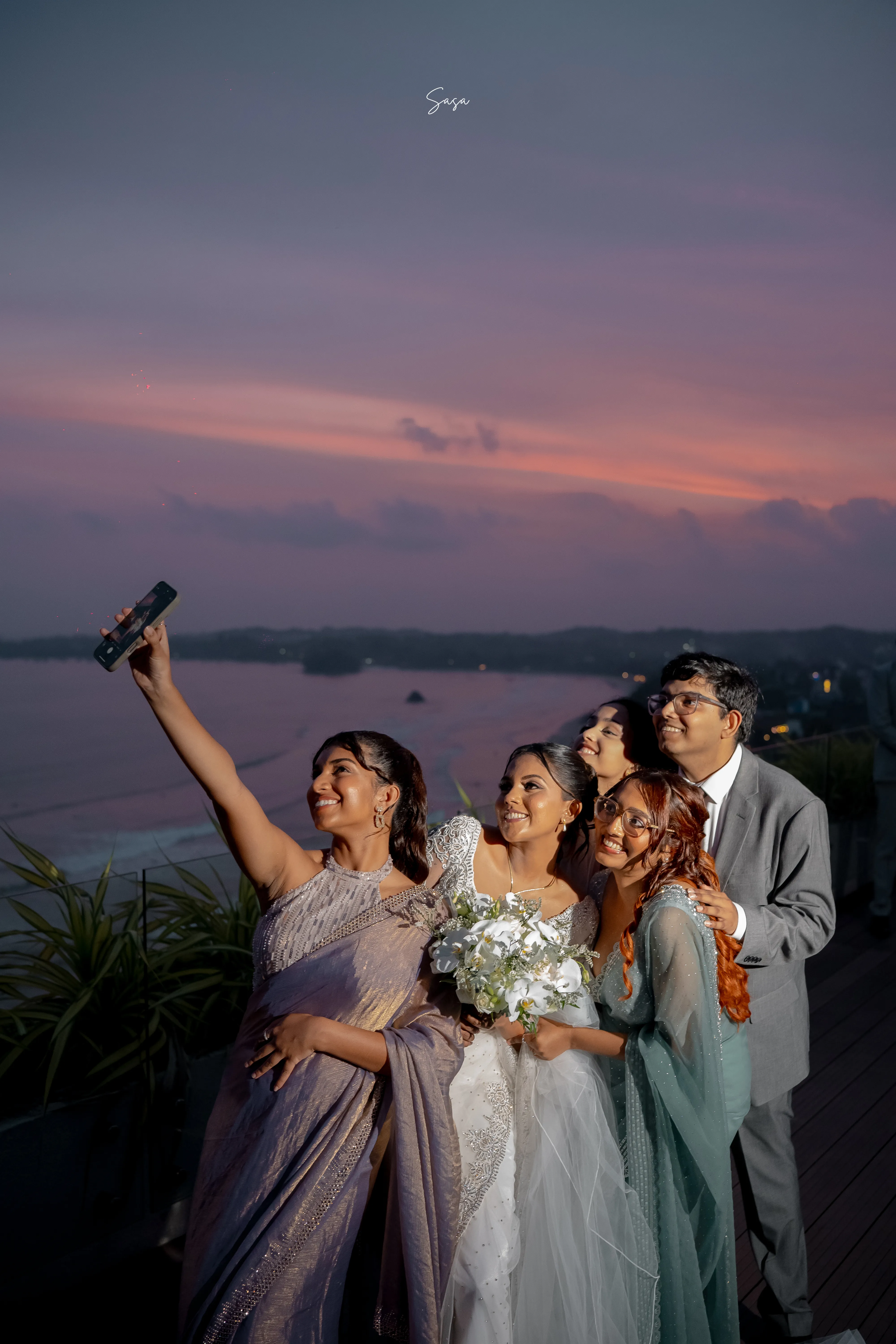 Harindi raises a phone for a cheerful group selfie with friends and family.