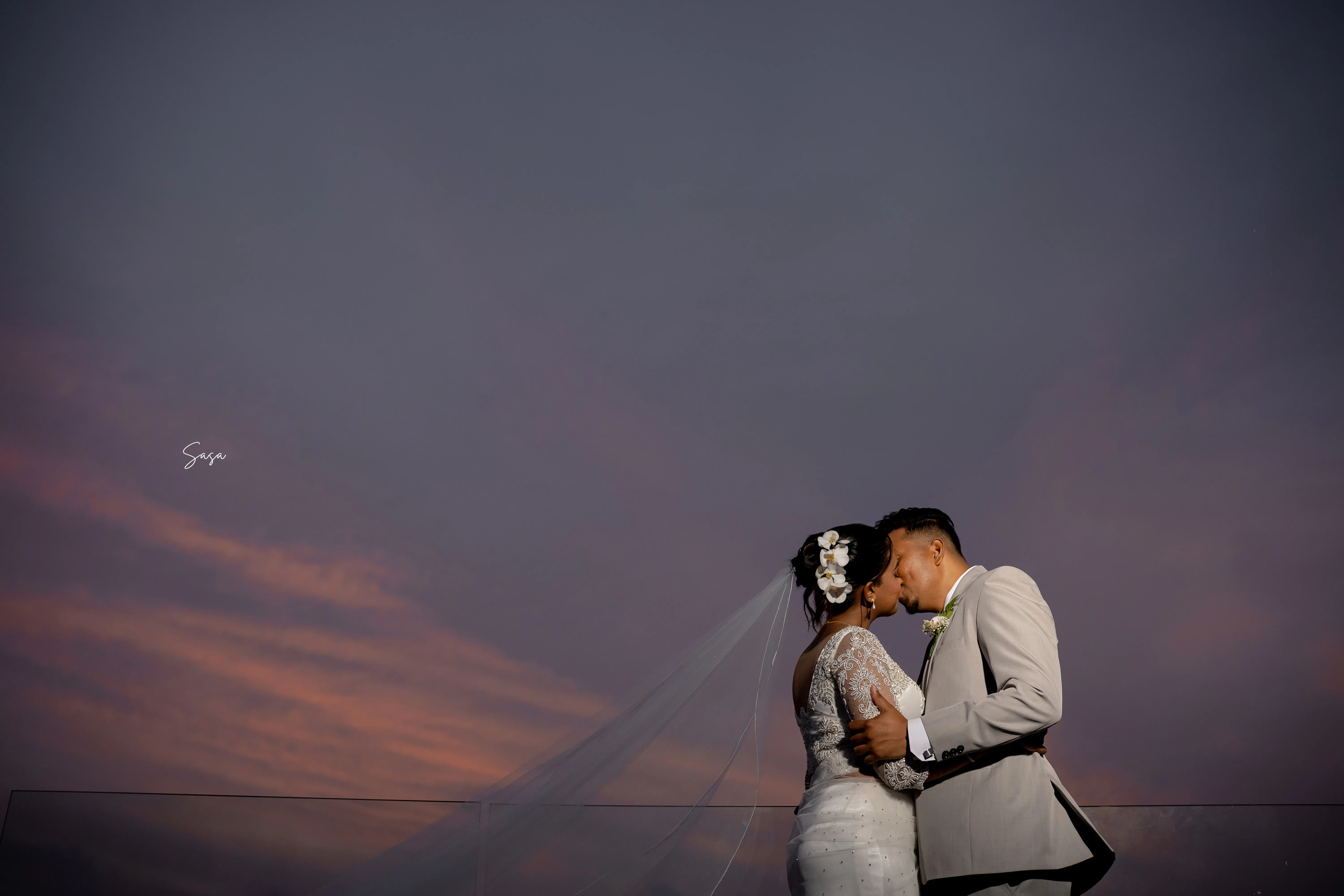 The couple shares a kiss beneath a violet sunset above the ocean.