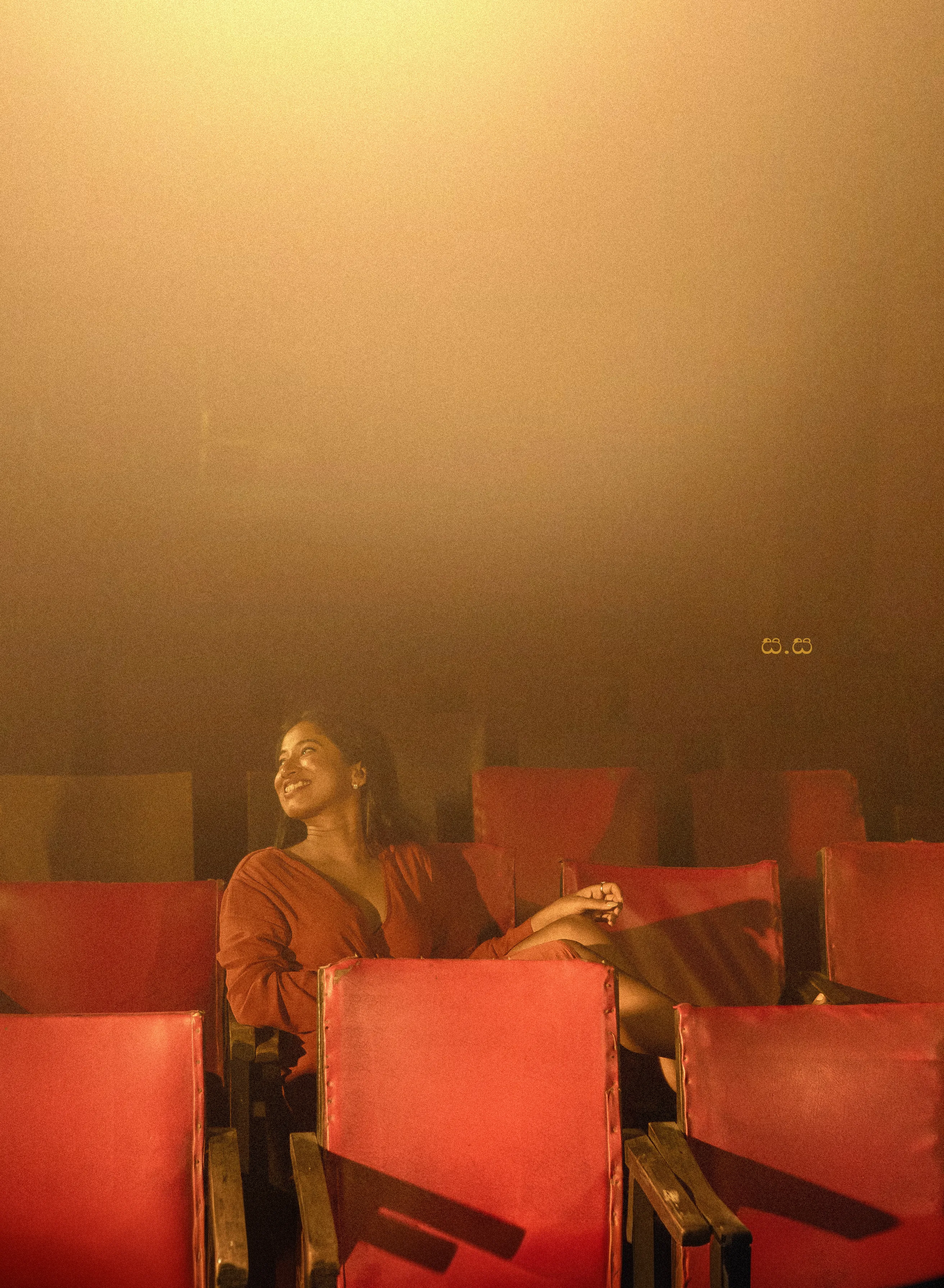 Hiruma reclines across vivid red seats in a warm theatrical portrait.