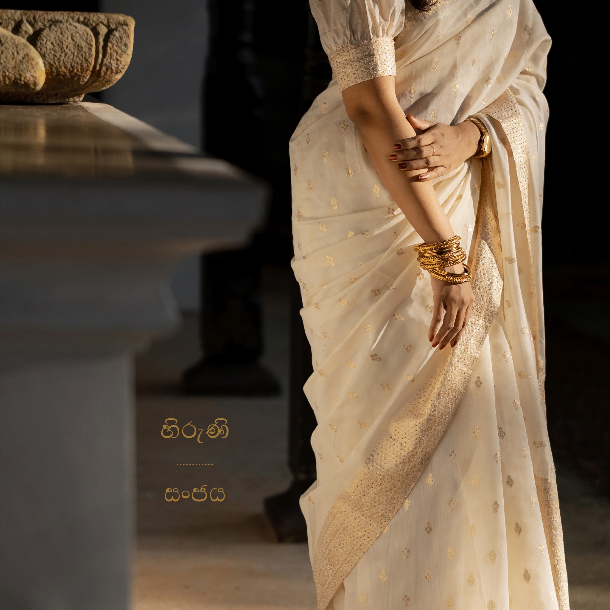A close crop highlights Hiruni’s ivory sari, gold bangles, and folded hands.