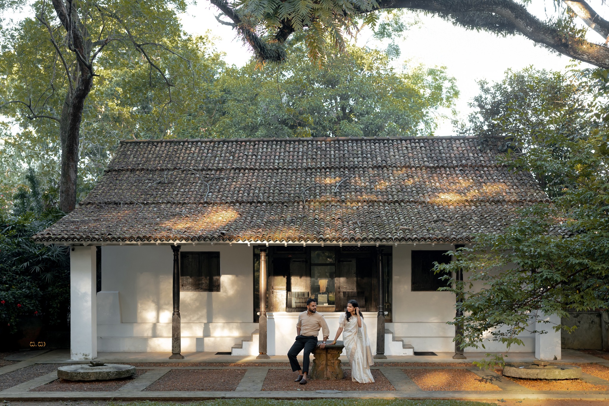 A wide portrait places the couple beneath a tiled roof and spreading garden tree.