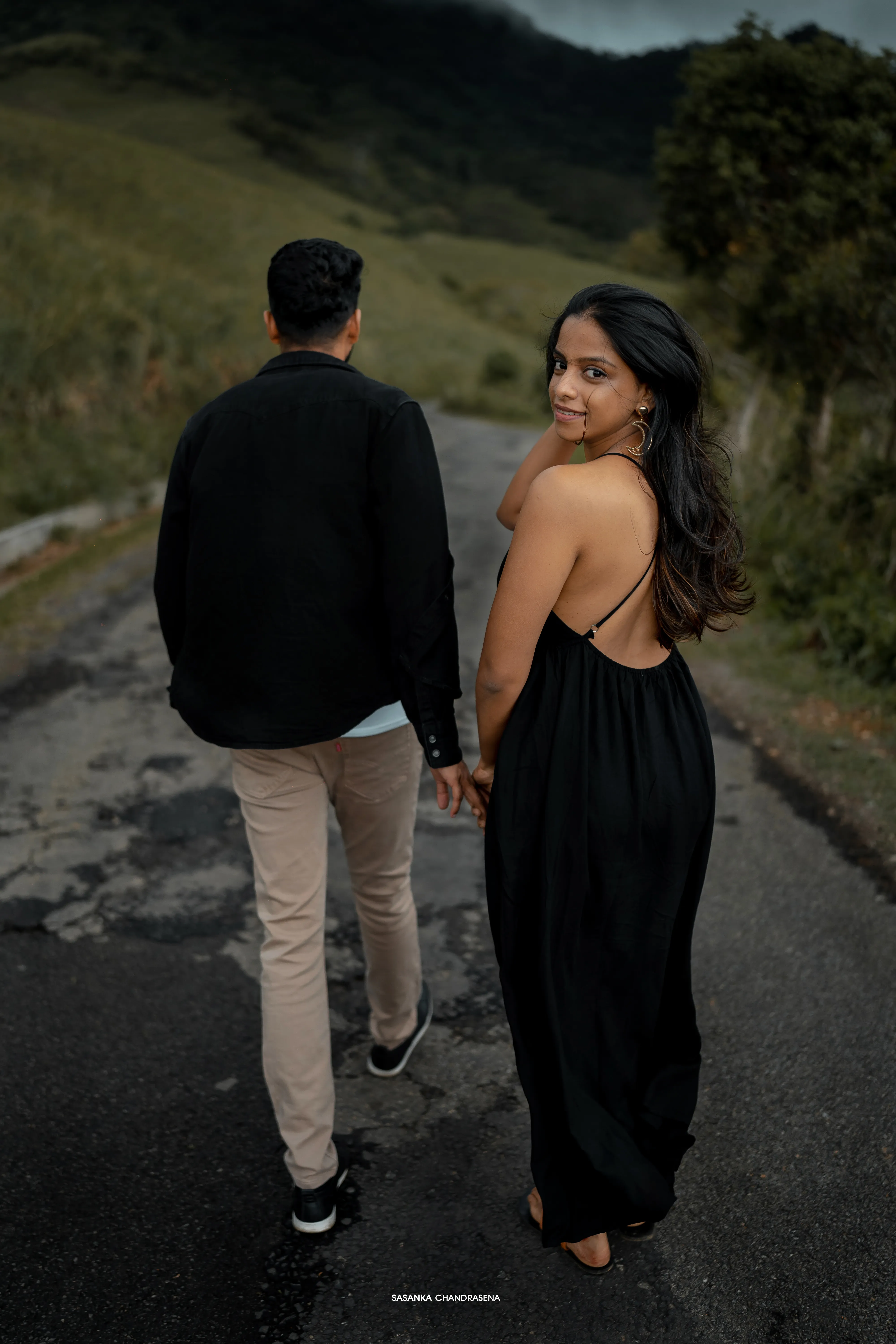 The couple holds hands while walking away along the mountain road.