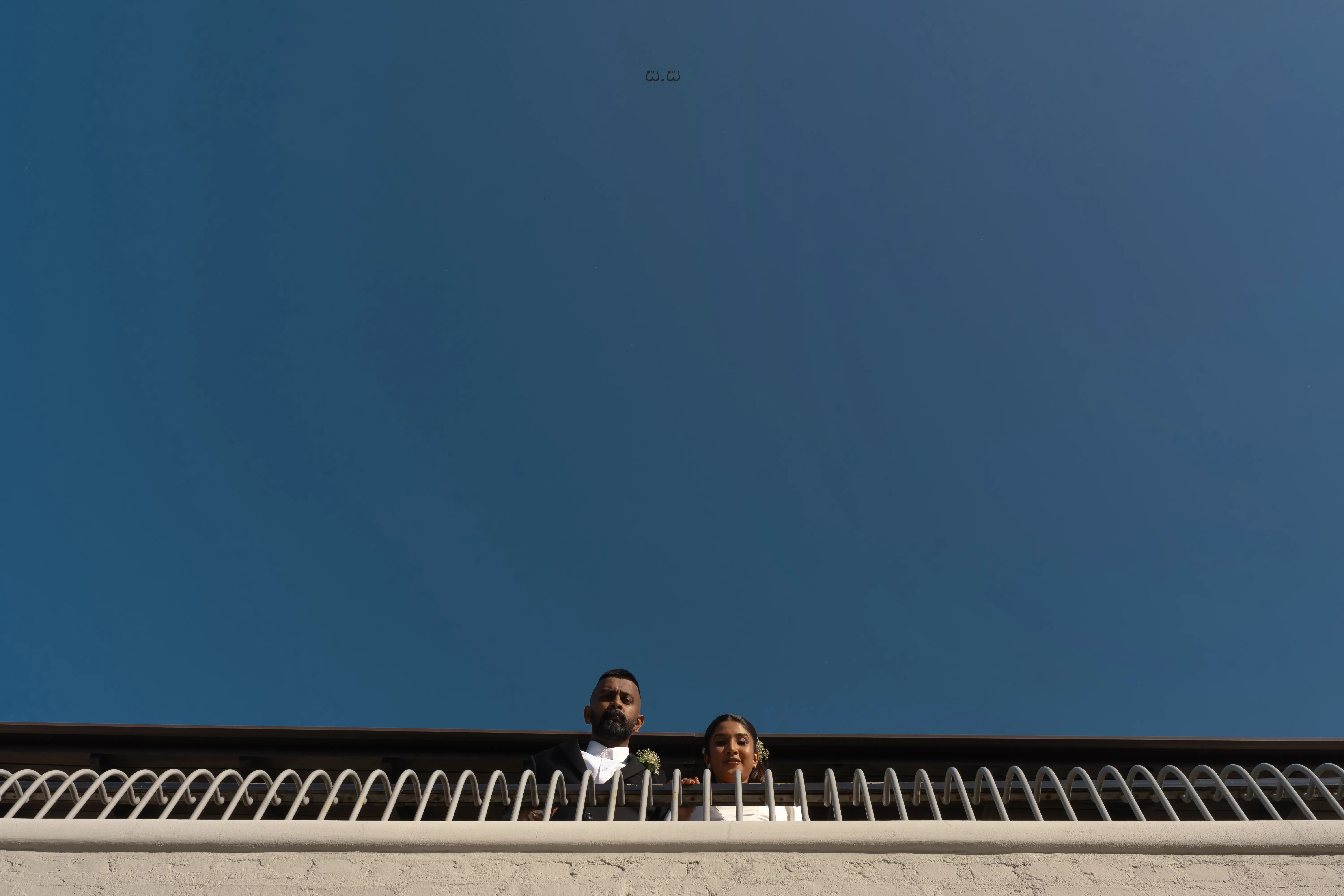 The couple looks over a white railing beneath an uninterrupted blue sky.