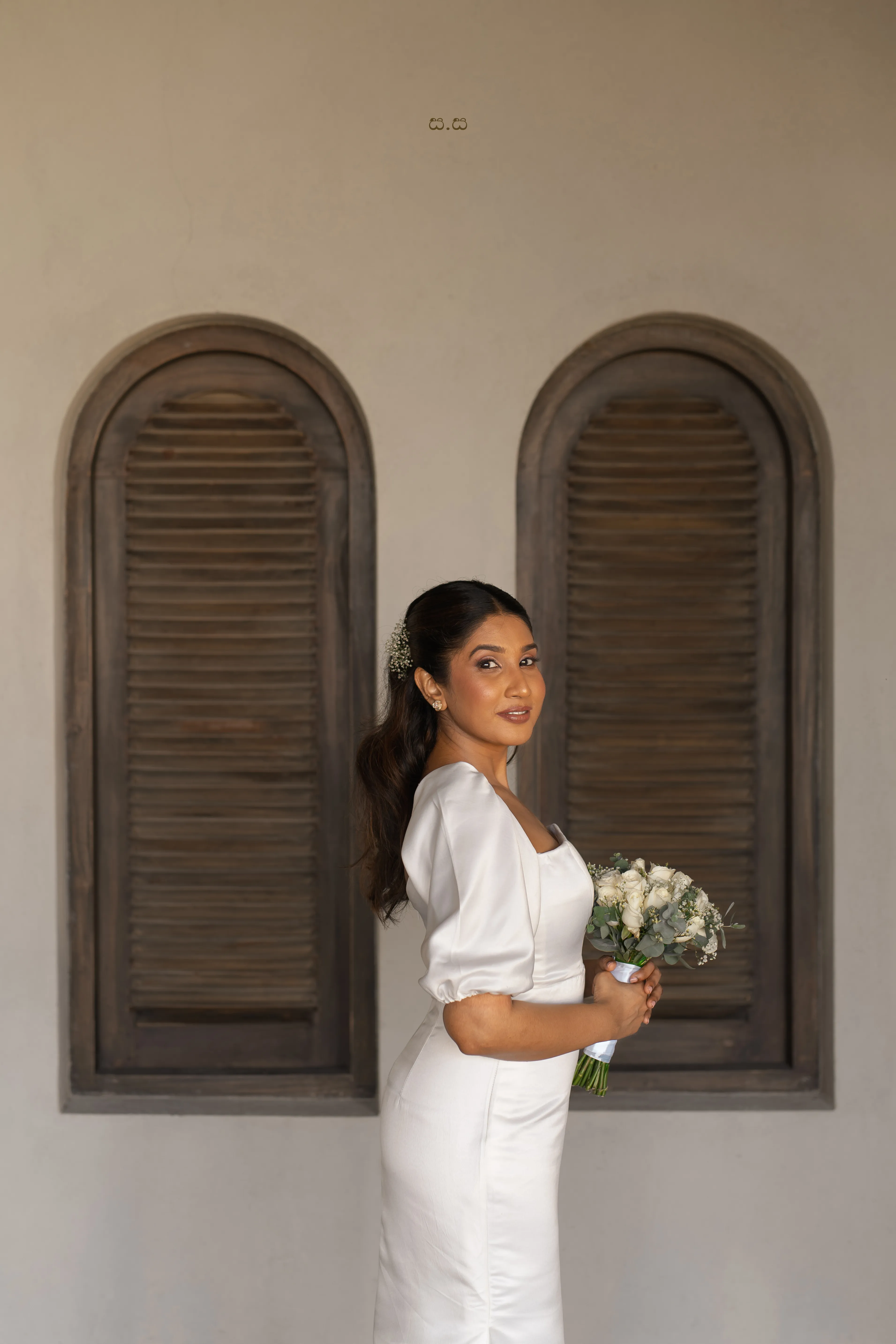 Malki holds a small white bouquet beside weathered wooden shutters.