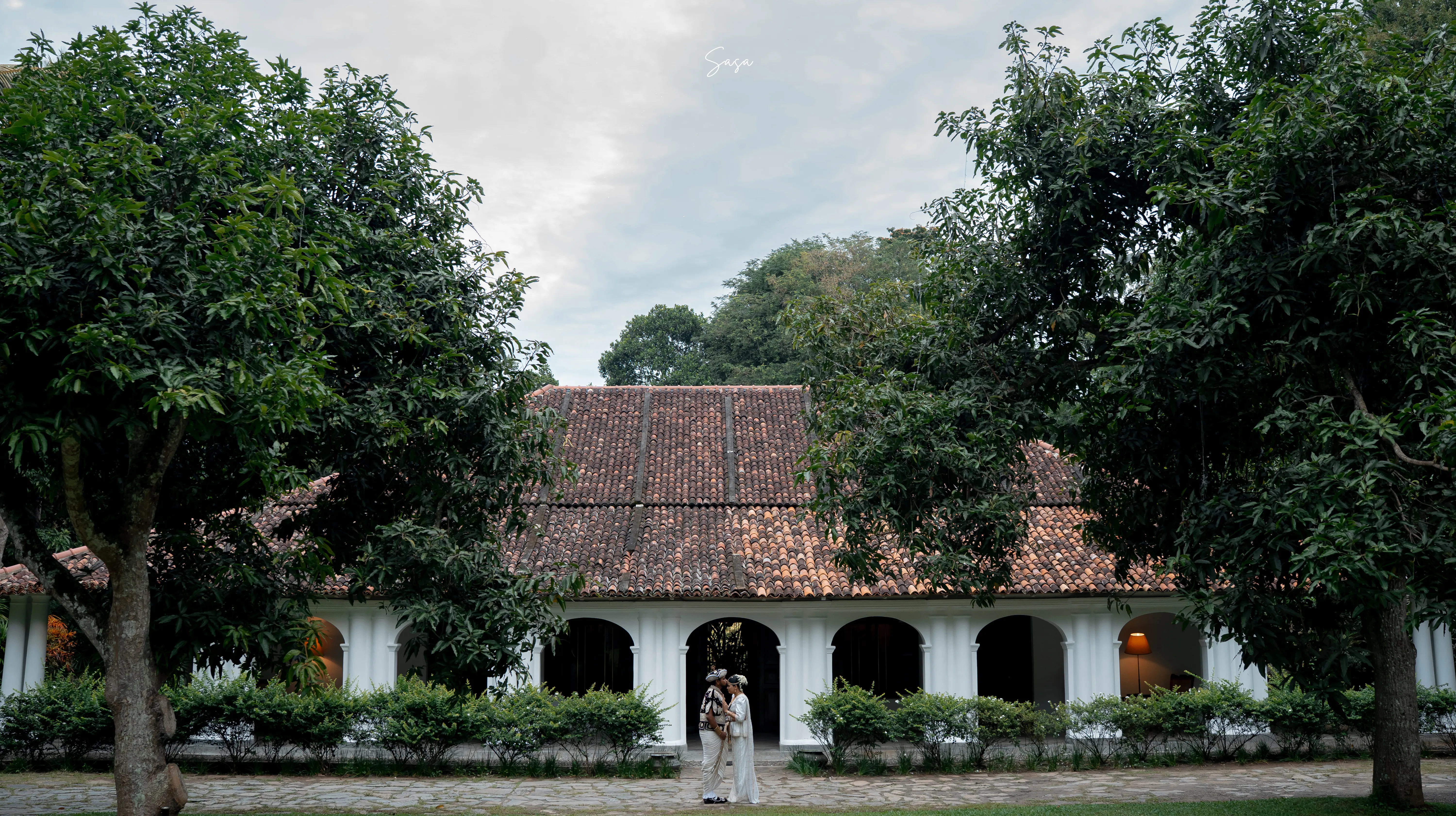 A wide portrait places the couple beneath the heritage house and mature trees.