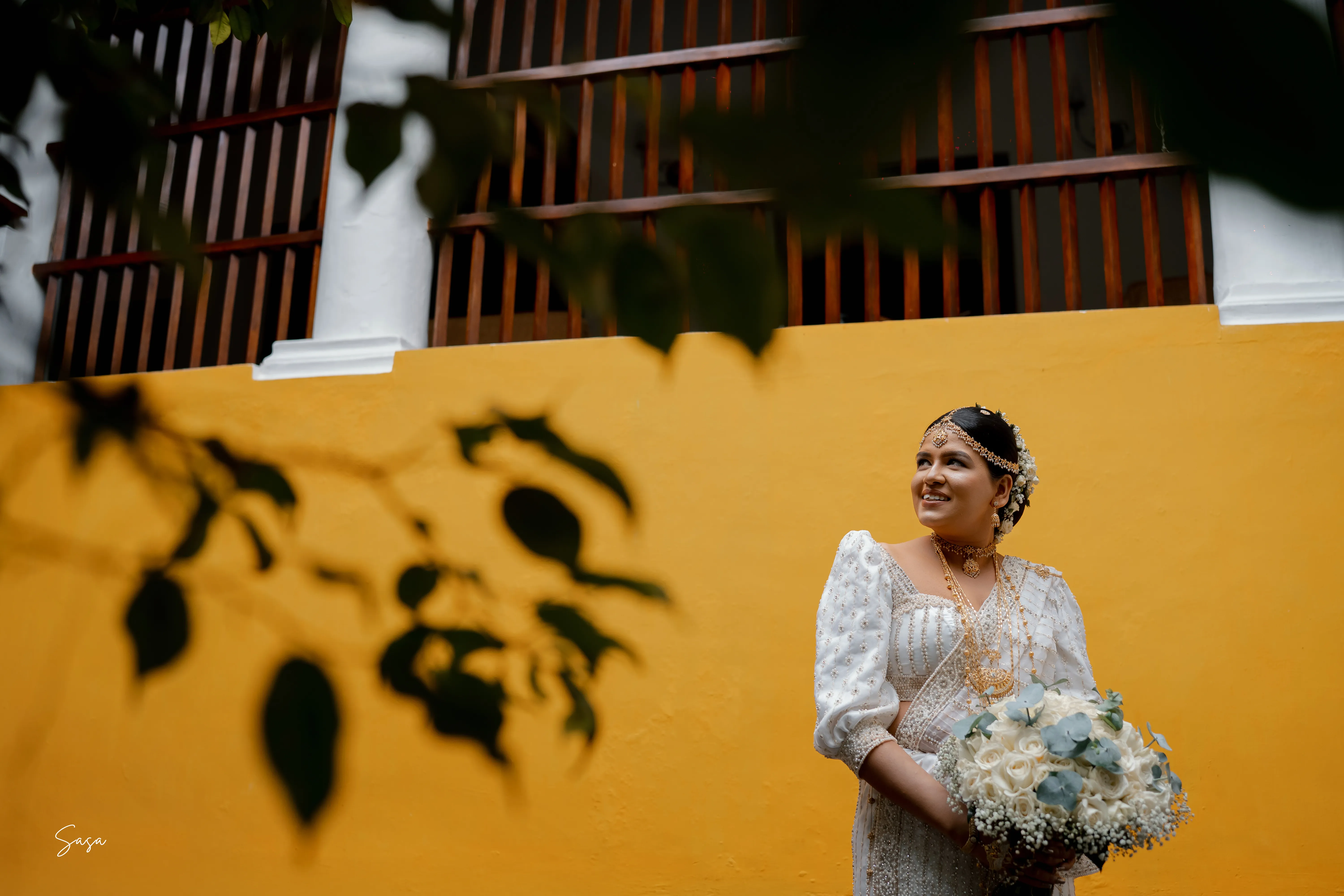 Nimasha holds her bouquet beside a sunlit ochre wall at the manor.