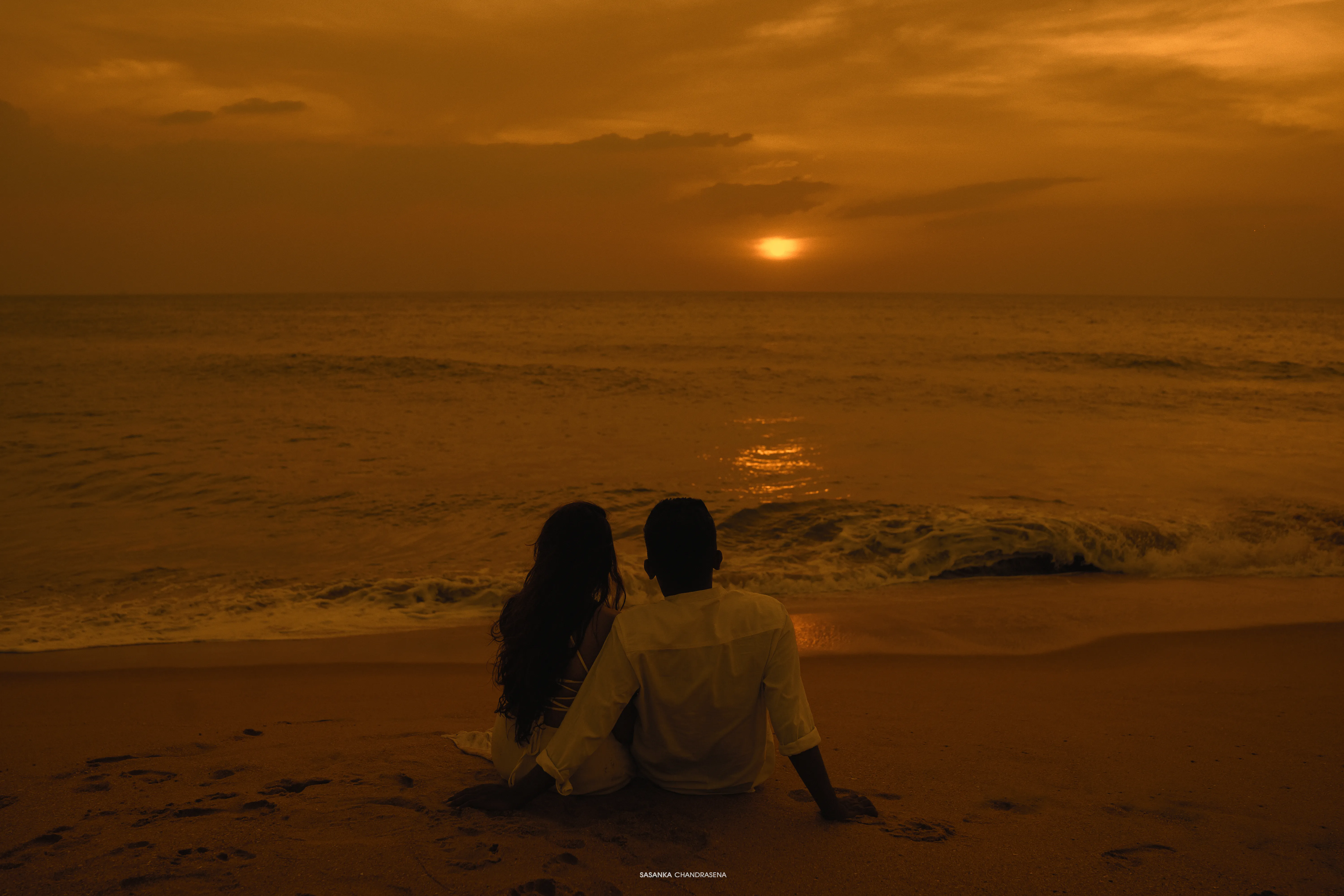 The couple sits together facing a deep amber sunset over the sea.