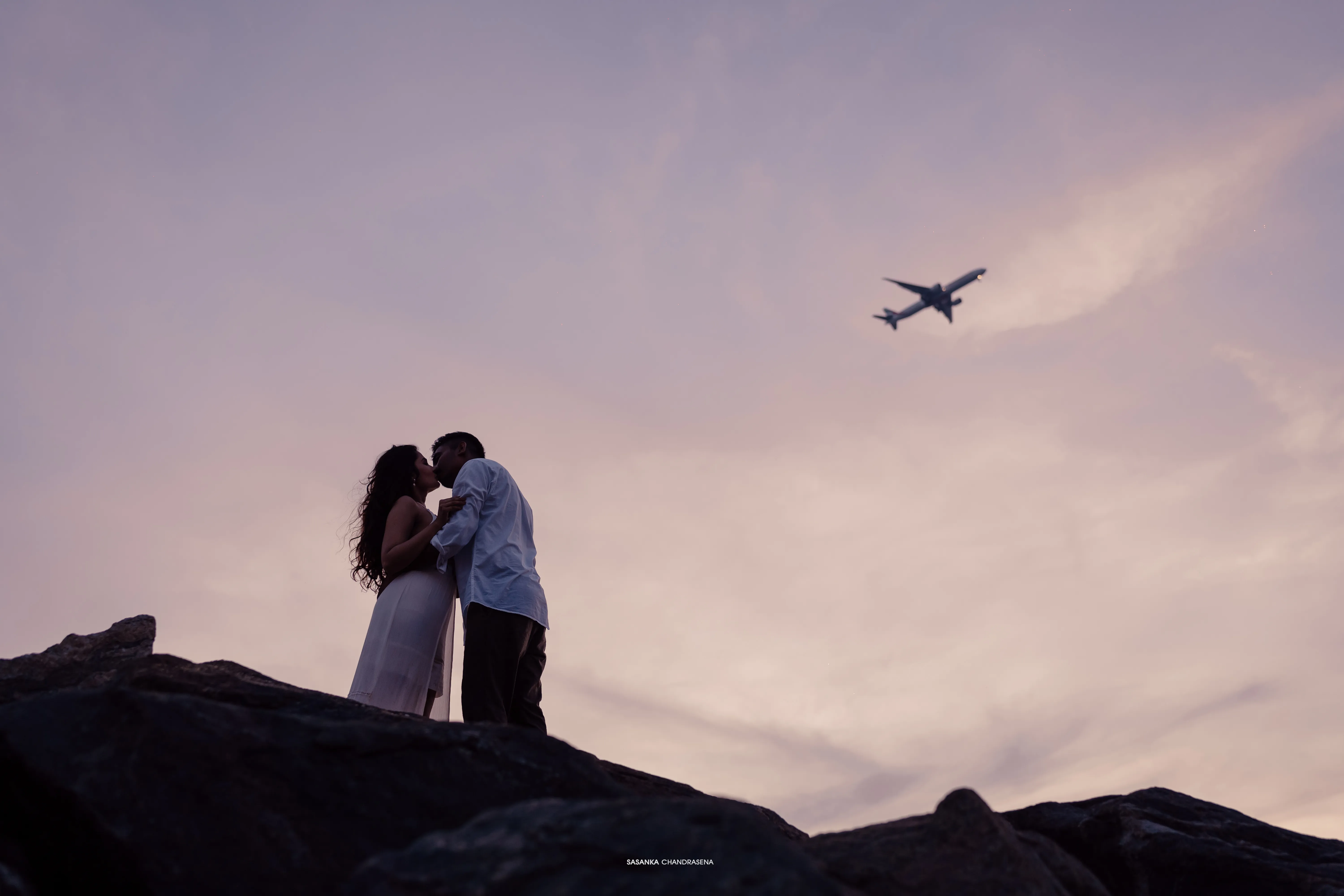 The couple meets on dark coastal rocks beneath the evening sky.