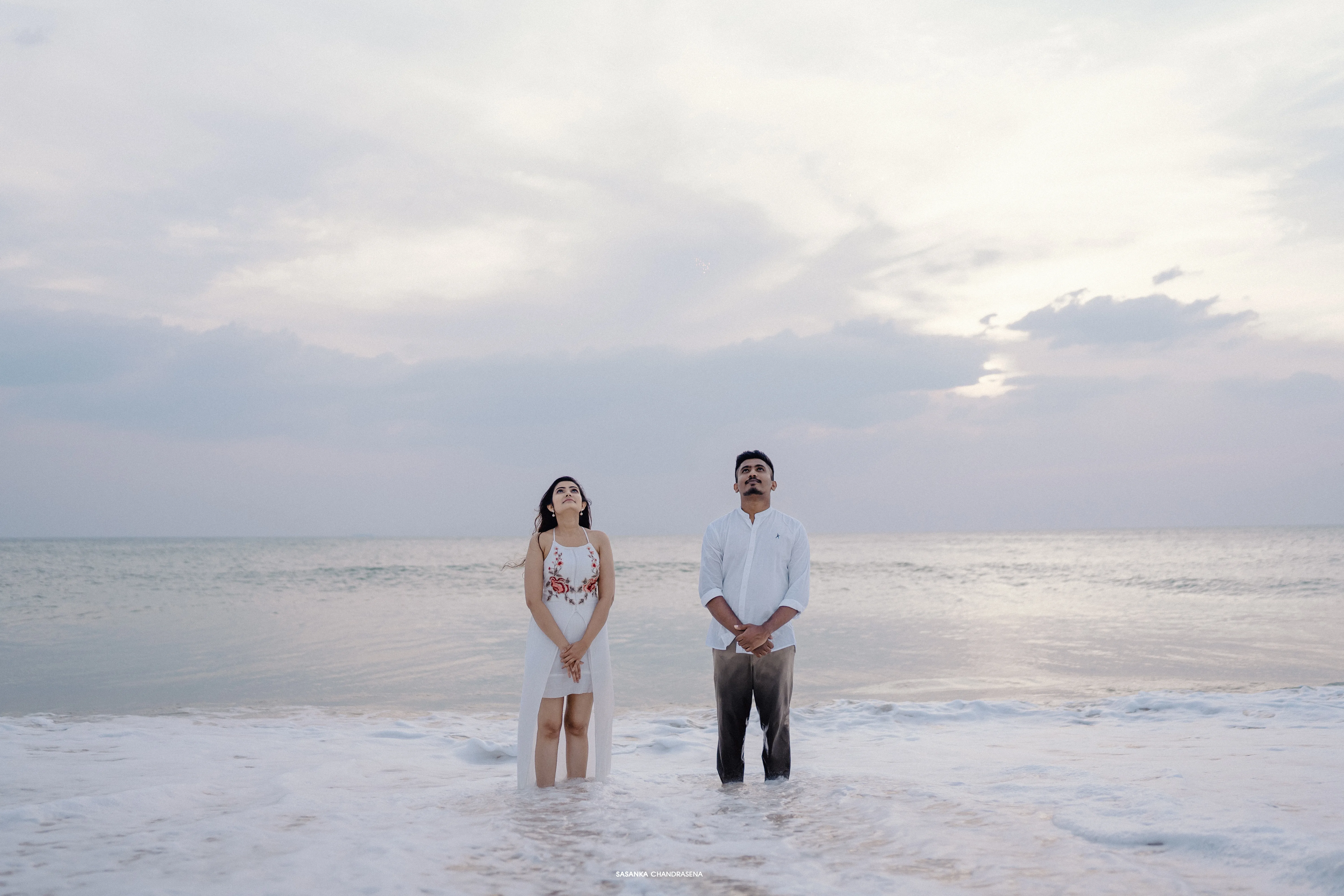 The couple faces the camera on a broad beach beneath soft evening clouds.