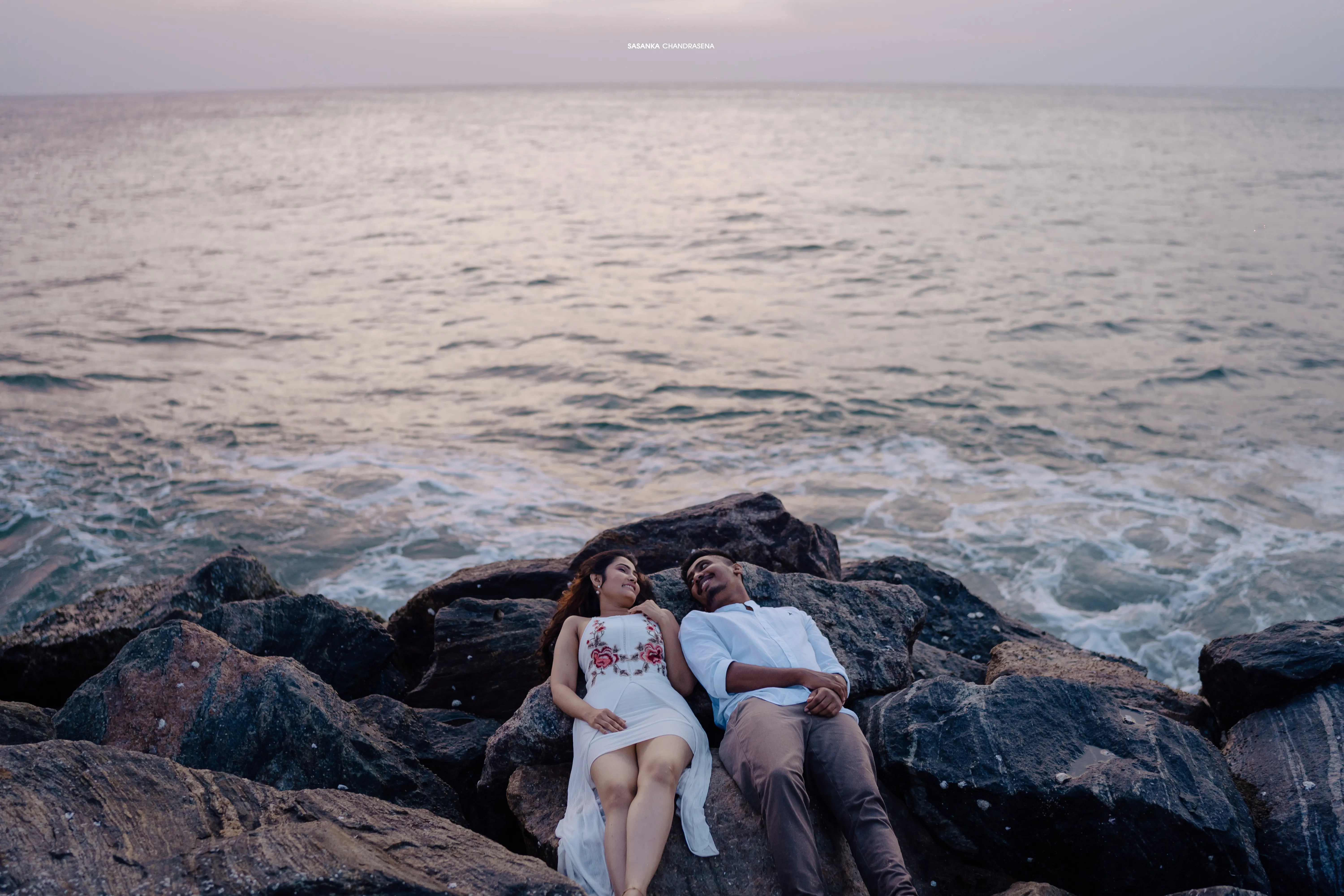 The couple sits together on dark rocks beside the moving sea.