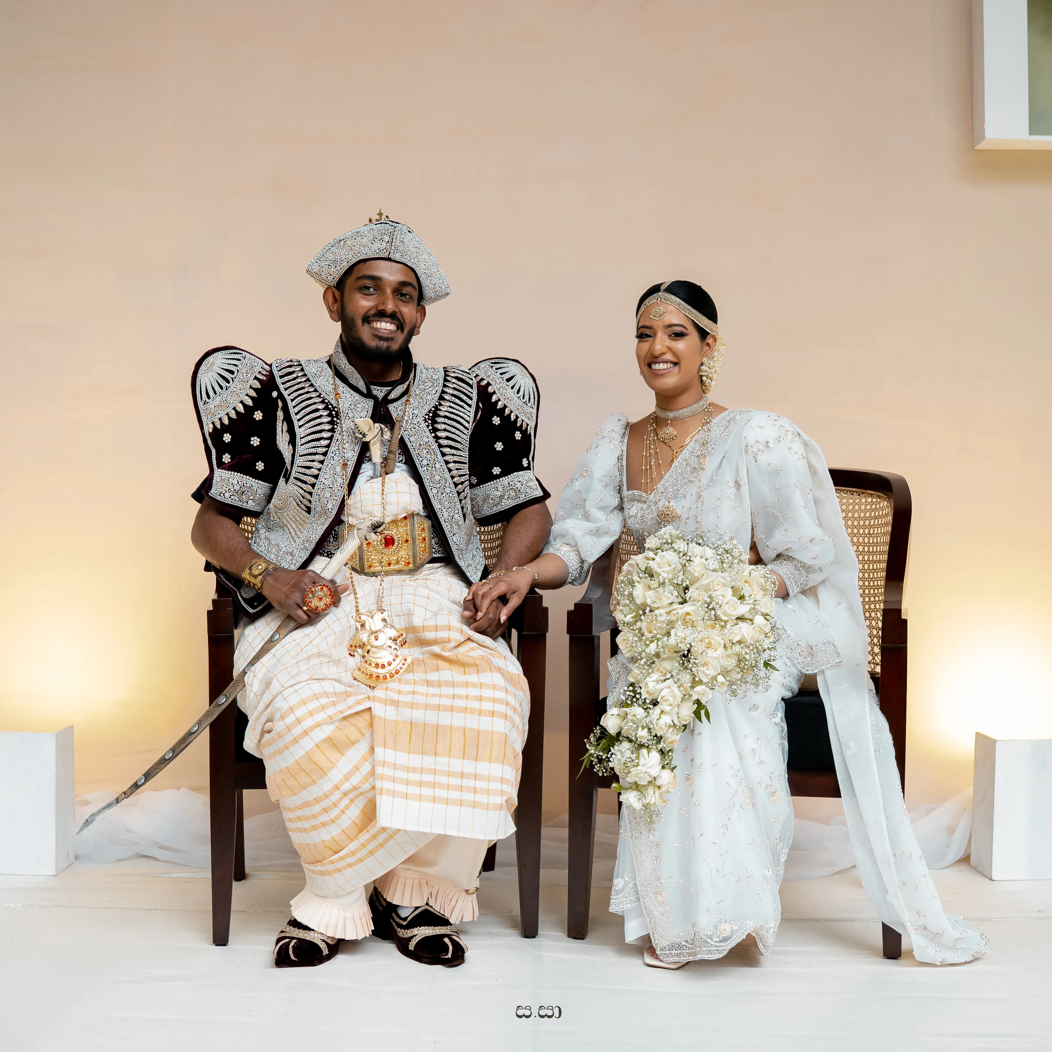 Shenuka and Sachini sit together in traditional ivory and embroidered attire.