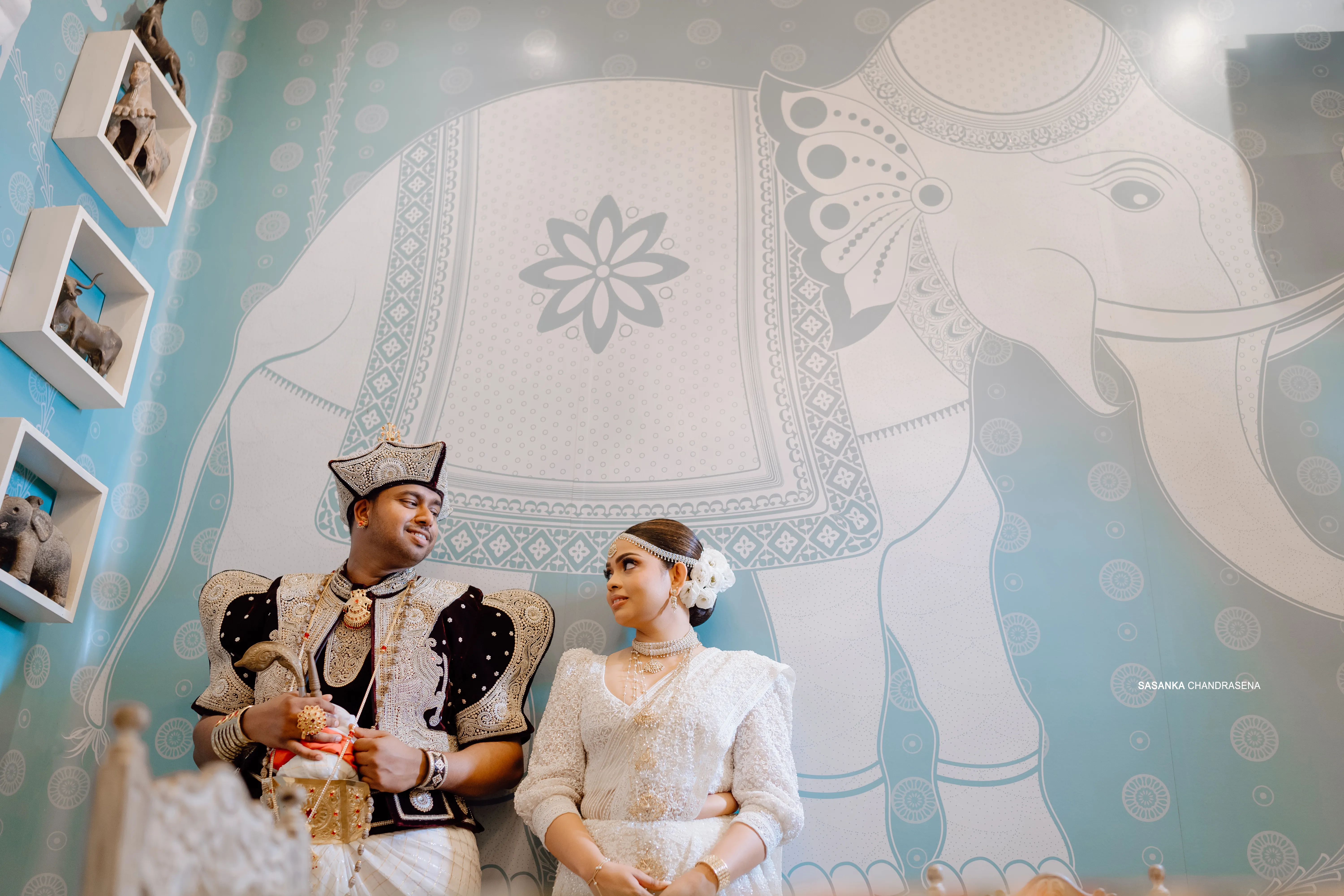 The newlyweds sit together against an ornate blue patterned wall.