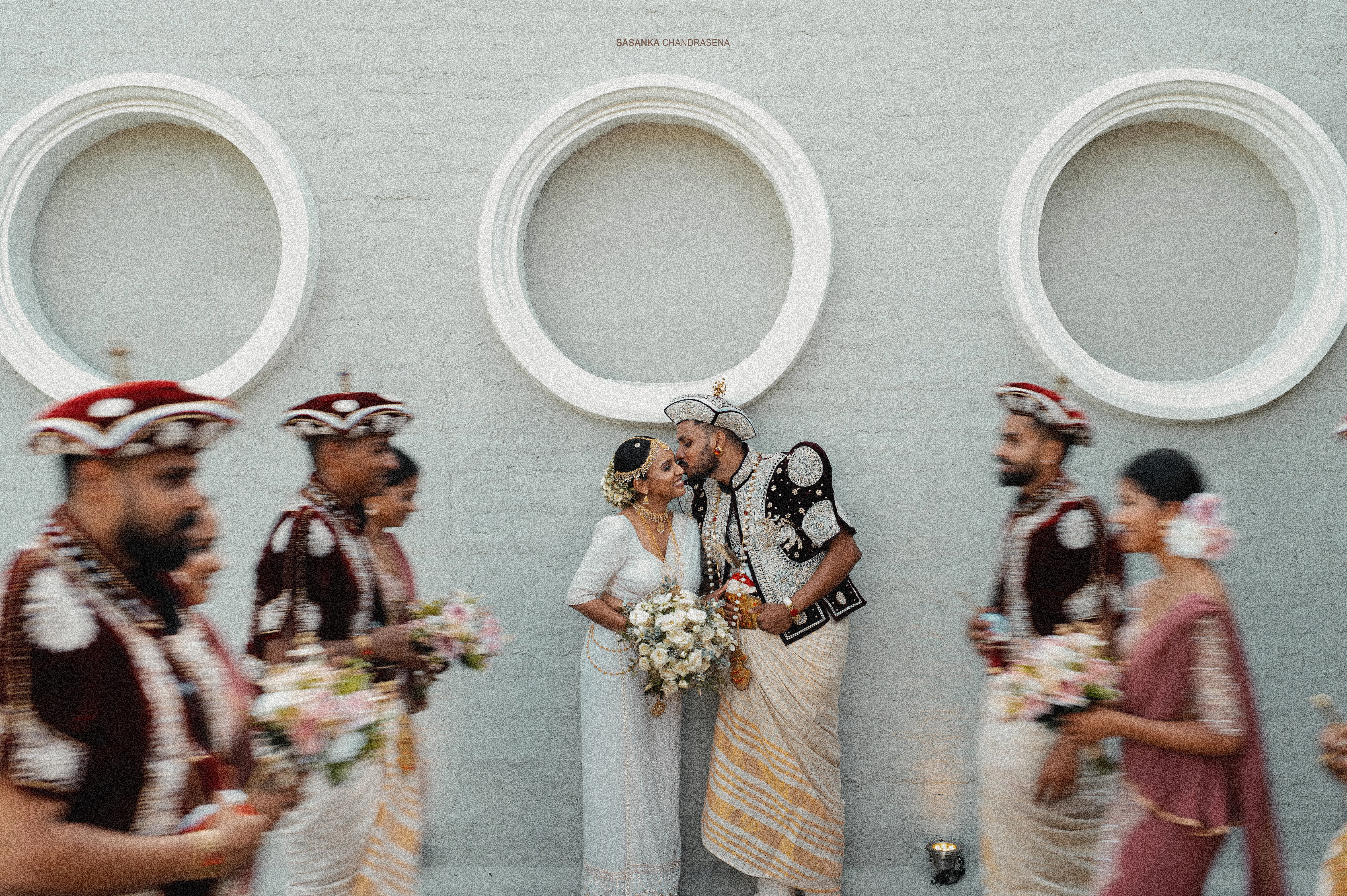 The couple stands together as the traditionally dressed wedding party passes through the frame.