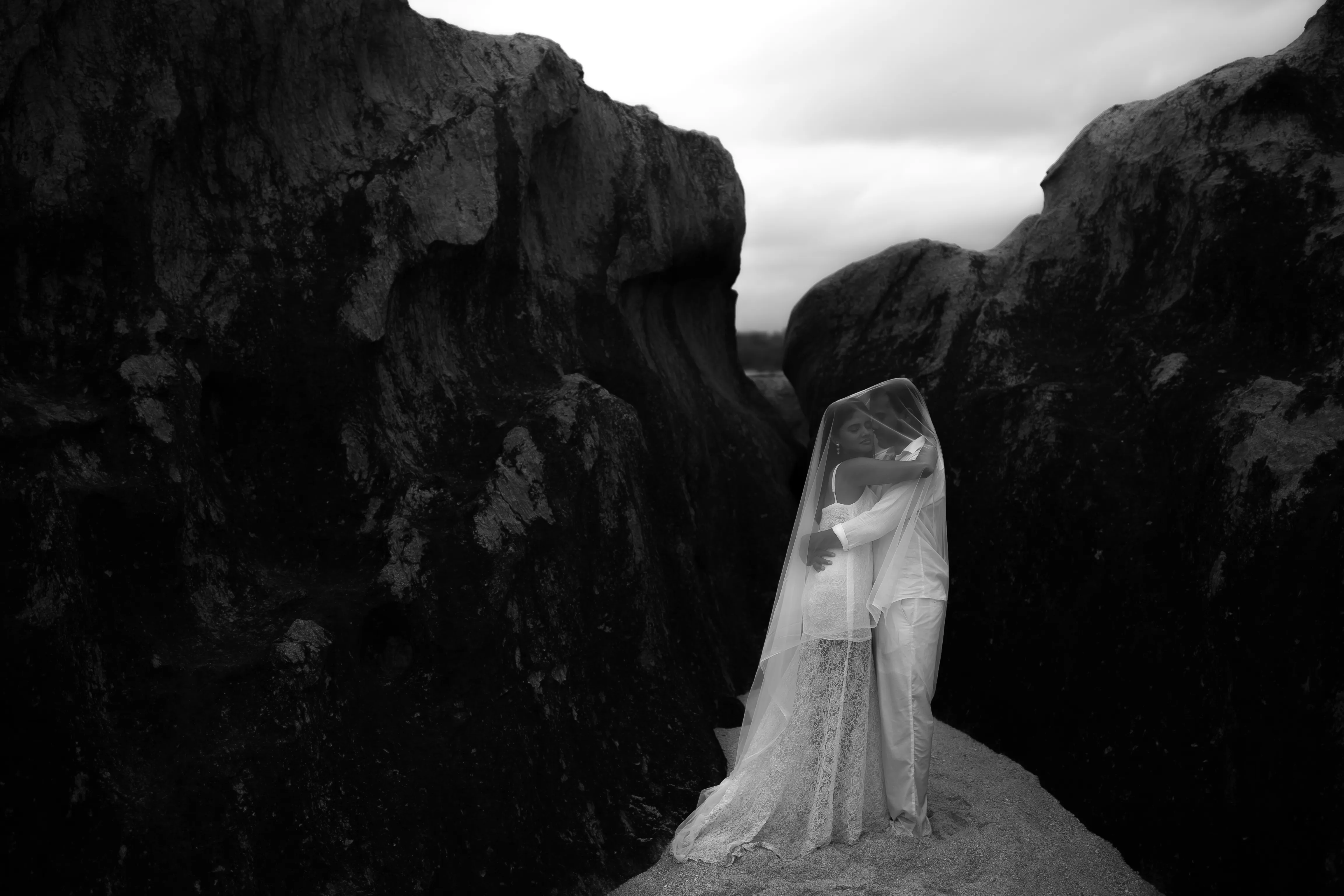 A black-and-white wedding portrait between dramatic coastal rocks