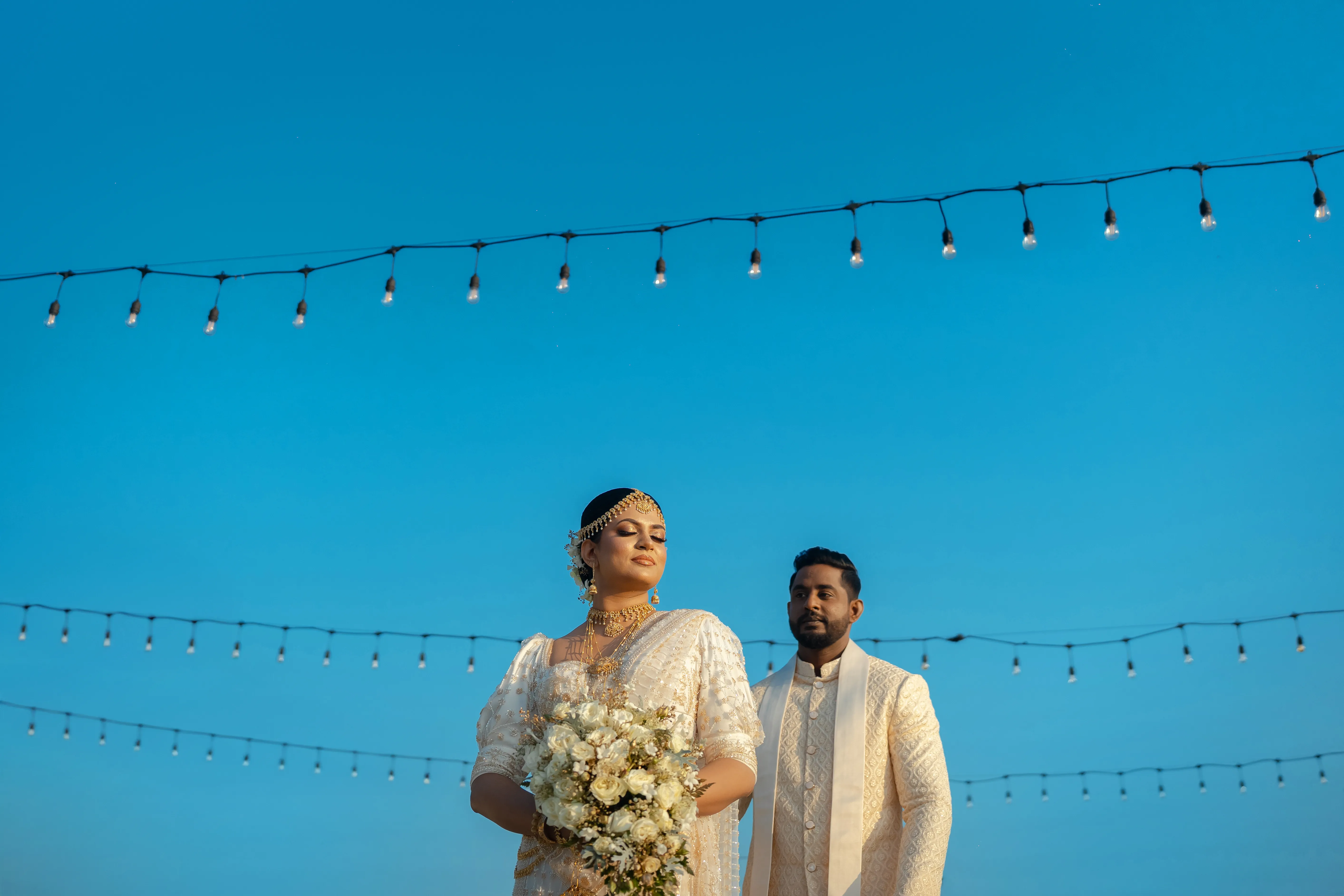 A Sri Lankan wedding couple beneath festoon lights and a clear blue sky